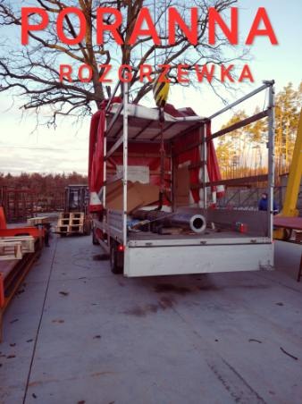 Ciężarówka z otwartą skrzynią załadowana rurami i elementami konstrukcyjnymi, w tle widać wózek widłowy i drzewa.