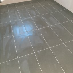 Stamarix - Świeżo położone, ciemnoszare płytki podłogowe z jasną fugą, widoczny fragment białej rury przy ścianie.