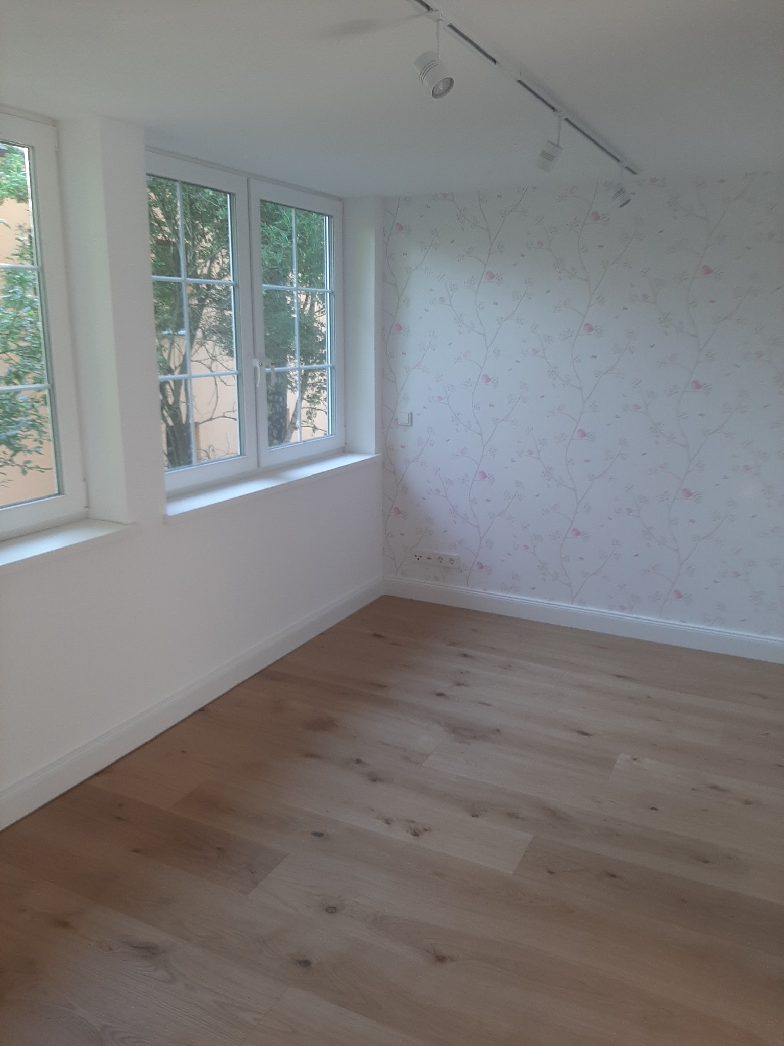 Pusta, jasna przestrzeń po remoncie z drewnianą podłogą, ścianą w delikatne kwiaty, dwoma oknami z widokiem na drzewa i oświetleniem sufitowym na szynie.