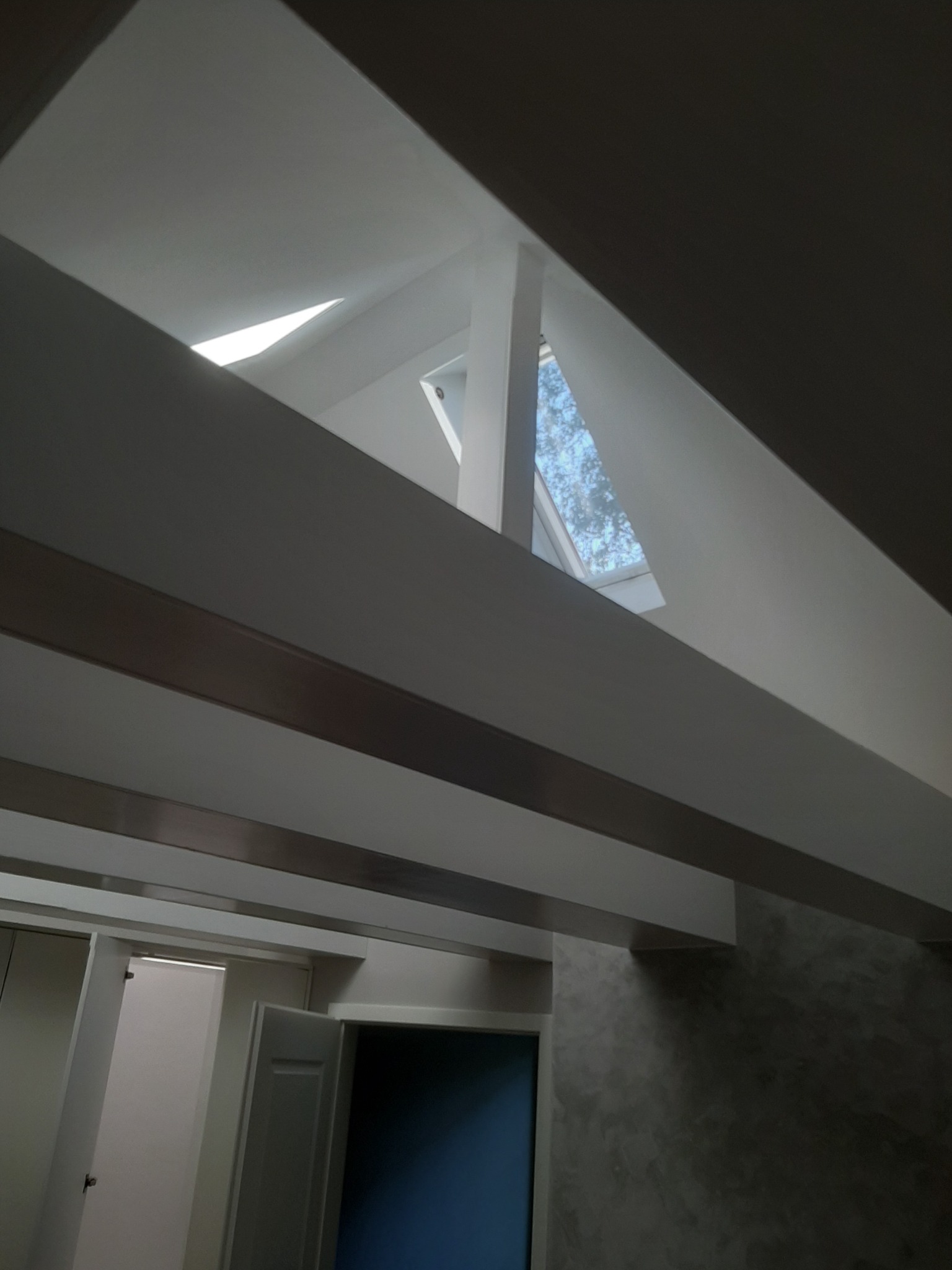 Wnętrze z widocznymi belkami sufitowymi, oknem dachowym i ścianą z efektem dekoracyjnym, widoczne fragmenty drzwi.