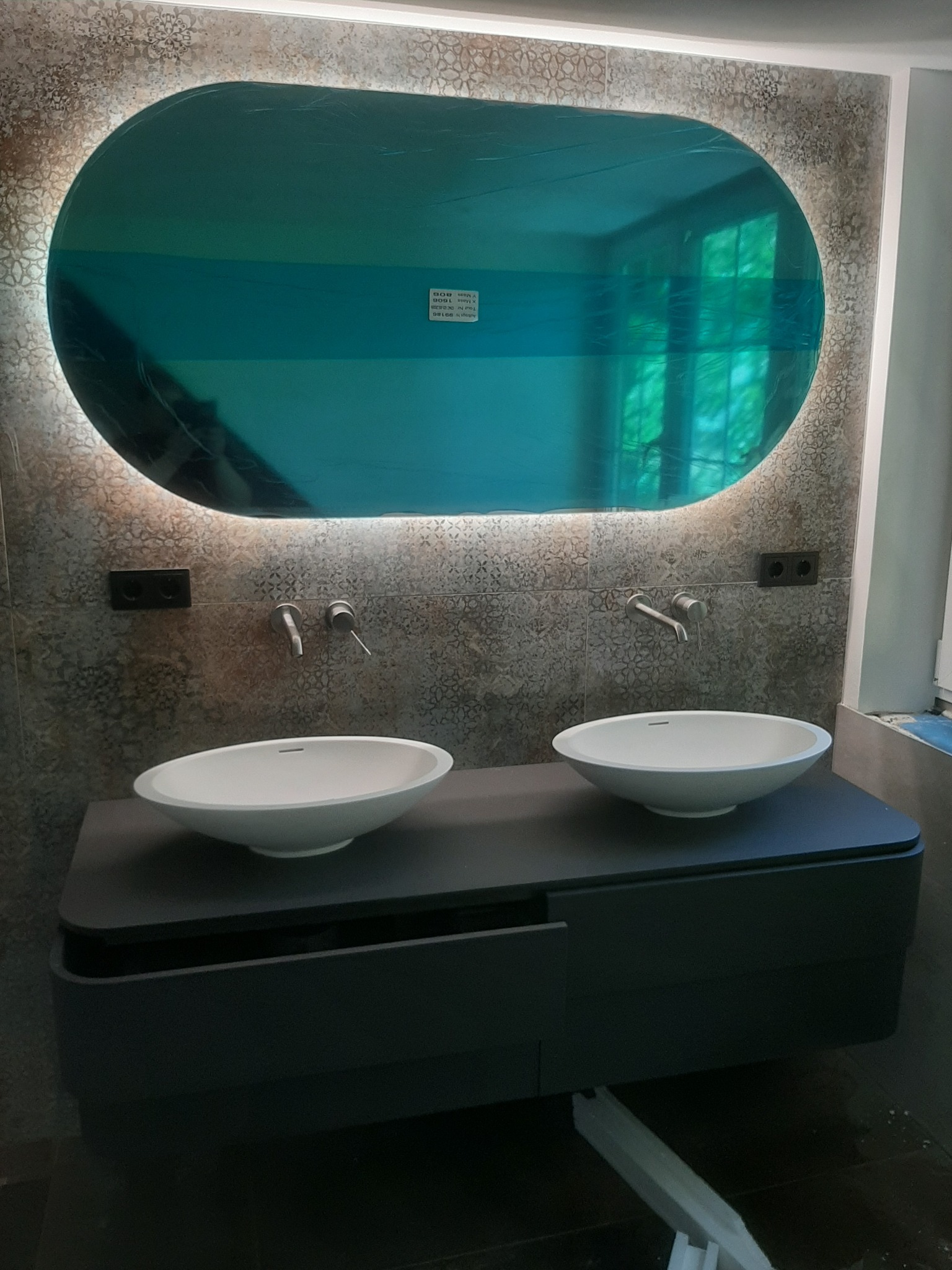 Nowoczesna łazienka z dwoma białymi umywalkami nablatowymi na czarnej szafce, owalne lustro z podświetleniem LED w folii ochronnej, szare płytki na ścianie