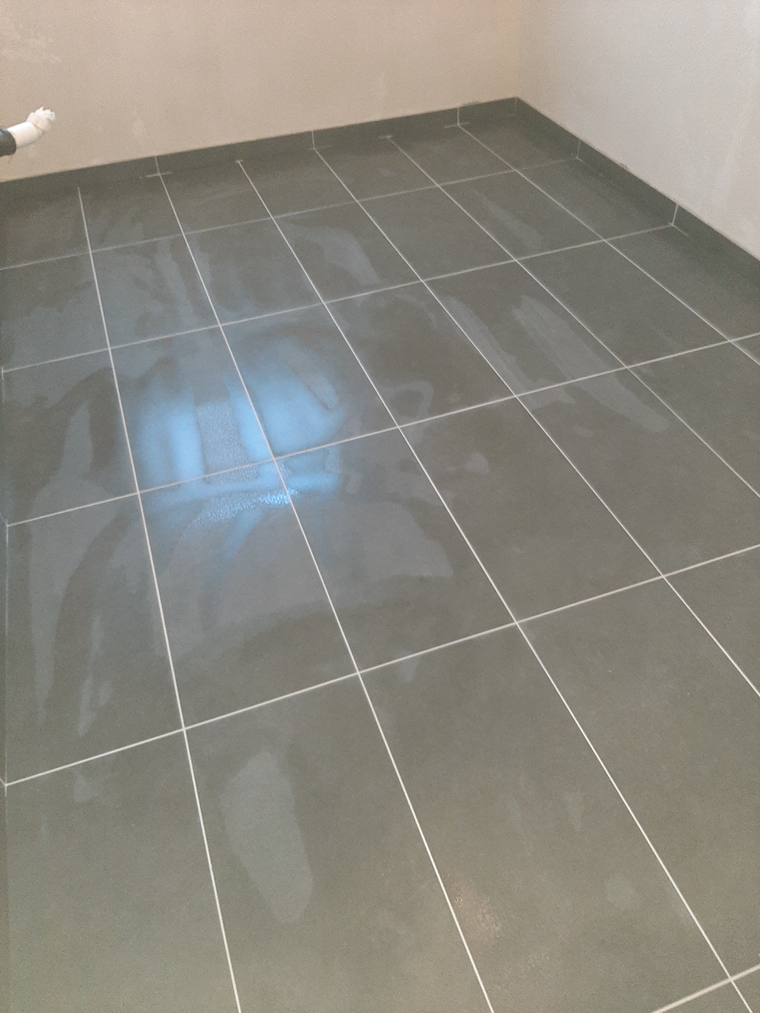 Świeżo położone, ciemnoszare płytki podłogowe z jasną fugą, widoczny fragment białej rury przy ścianie.