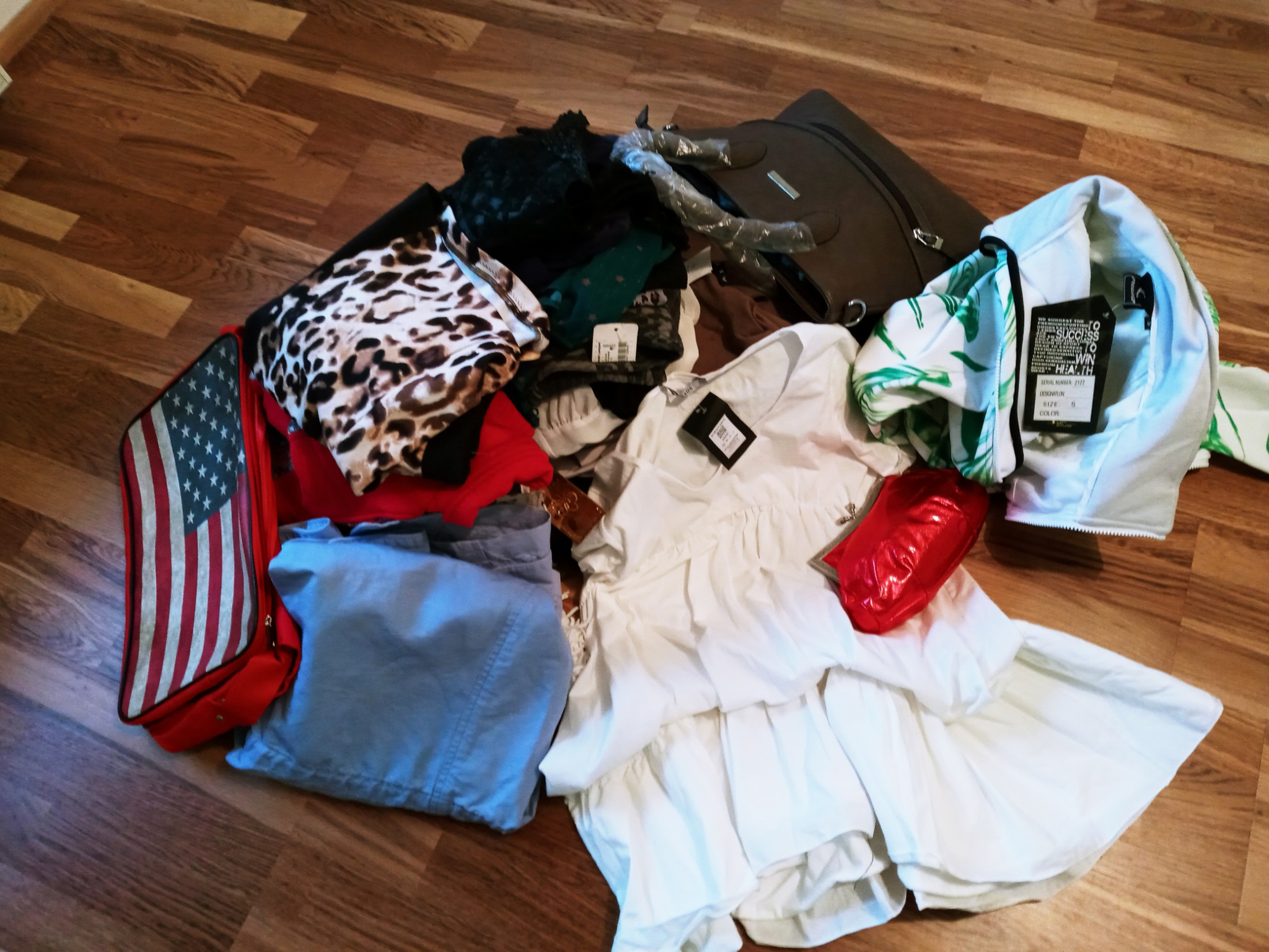 Stos ubrań damskich i męskich z metkami, torba podróżna i torba w barwach flagi USA na podłodze z paneli.