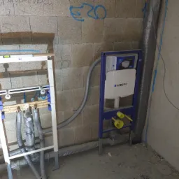 Stelaże podtynkowe Geberit zamontowane przy surowej ścianie z betonu komórkowego z widocznymi przewodami kanalizacyjnymi i wodnymi w trakcie instalacji.