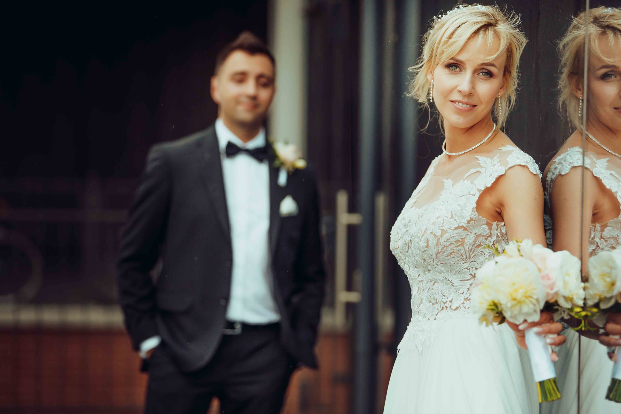 Panna młoda w koronkowej sukni ślubnej, trzymająca bukiet z białych i różowych kwiatów, odbija się w lustrze, z uśmiechem spogląda w obiektyw, w tle rozmyty pan młody w czarnym smokingu.