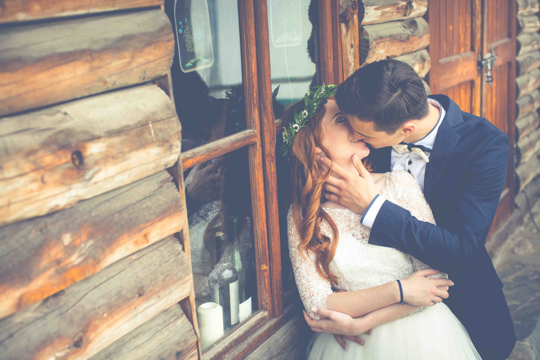 Para młoda w objęciach, on w granatowym garniturze z muszką, ona w koronkowej sukni i wianku, całują się na tle drewnianej ściany z oknem, w którym widać butelki i świecę.