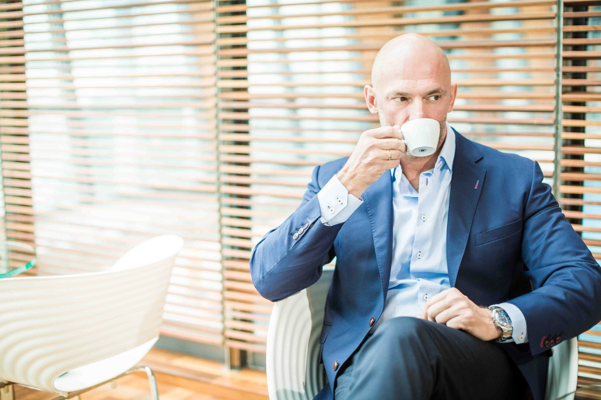 Łysy mężczyzna w niebieskiej marynarce pije kawę w nowoczesnym biurze z drewnianymi żaluzjami.