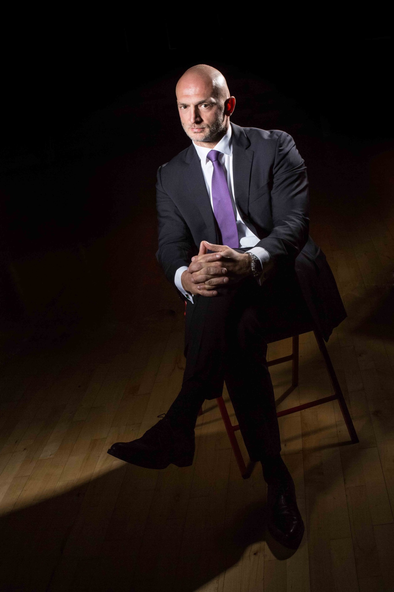 Portret mężczyzny w garniturze i fioletowym krawacie, siedzącego na krześle w ciemnym pomieszczeniu, oświetlonego punktowym światłem.