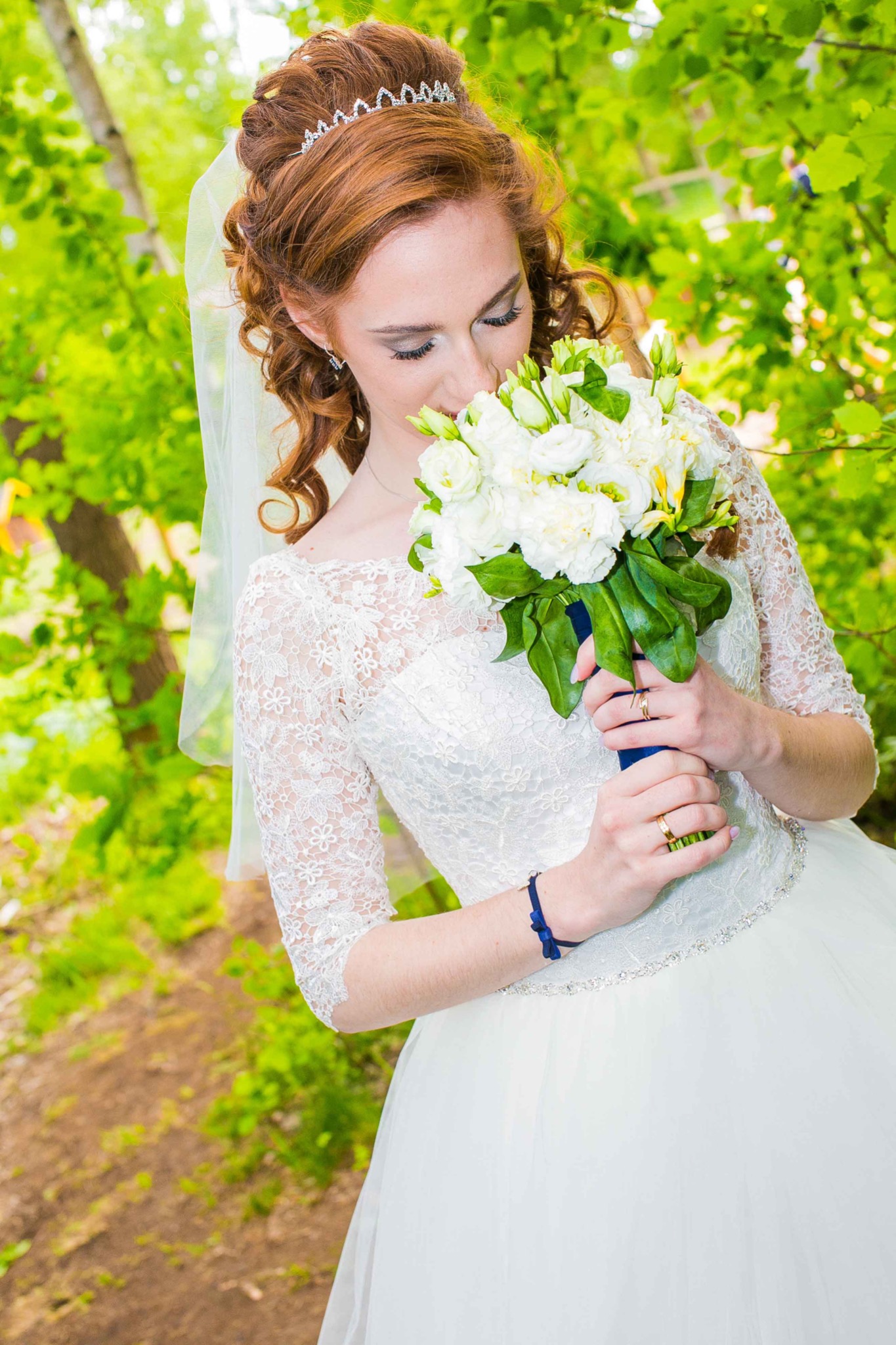 Panna młoda w koronkowej sukni ślubnej z tiulowym welonem i diademem, wącha bukiet białych kwiatów na tle zieleni.