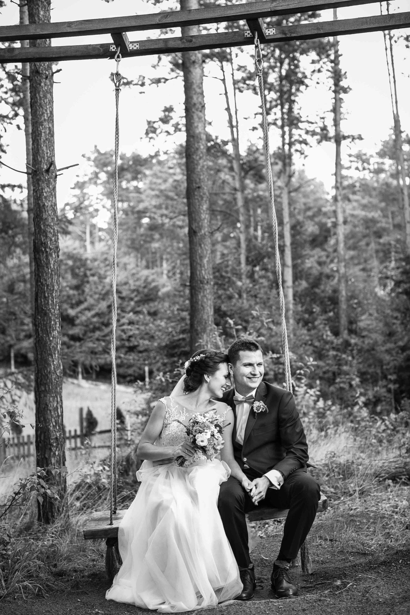 Czarno-białe zdjęcie uśmiechniętej pary młodej siedzącej na huśtawce z desek i liny w lesie, panna młoda w sukni z koronkową górą trzyma bukiet, pan młody w ciemnym garniturze z muszką.