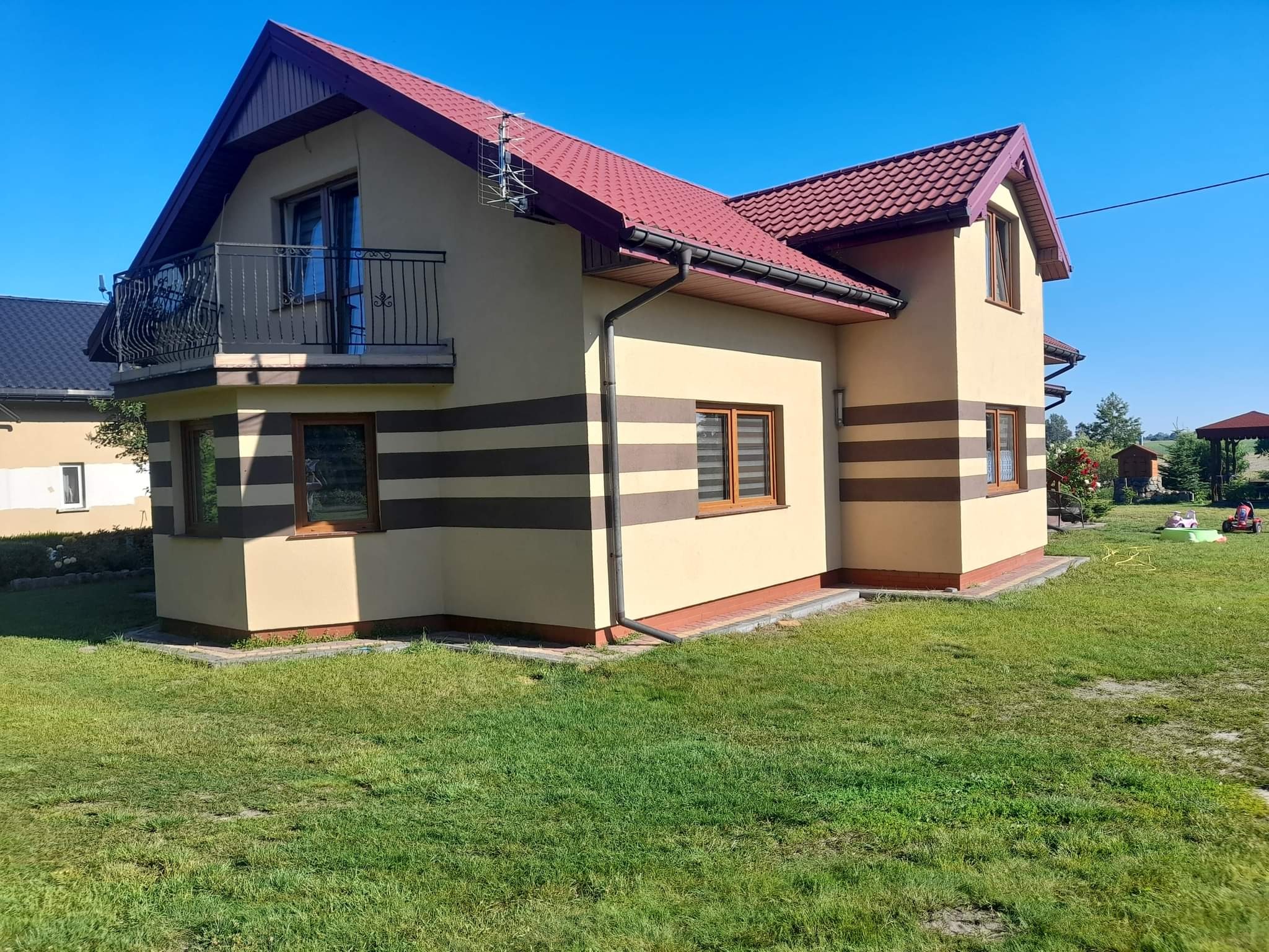Elewacja domu jednorodzinnego w odcieniach beżu z poziomymi pasami w kolorze brązowym, czerwony dach z metalową anteną, balkon z ozdobną balustradą, widok z poziomu trawnika.