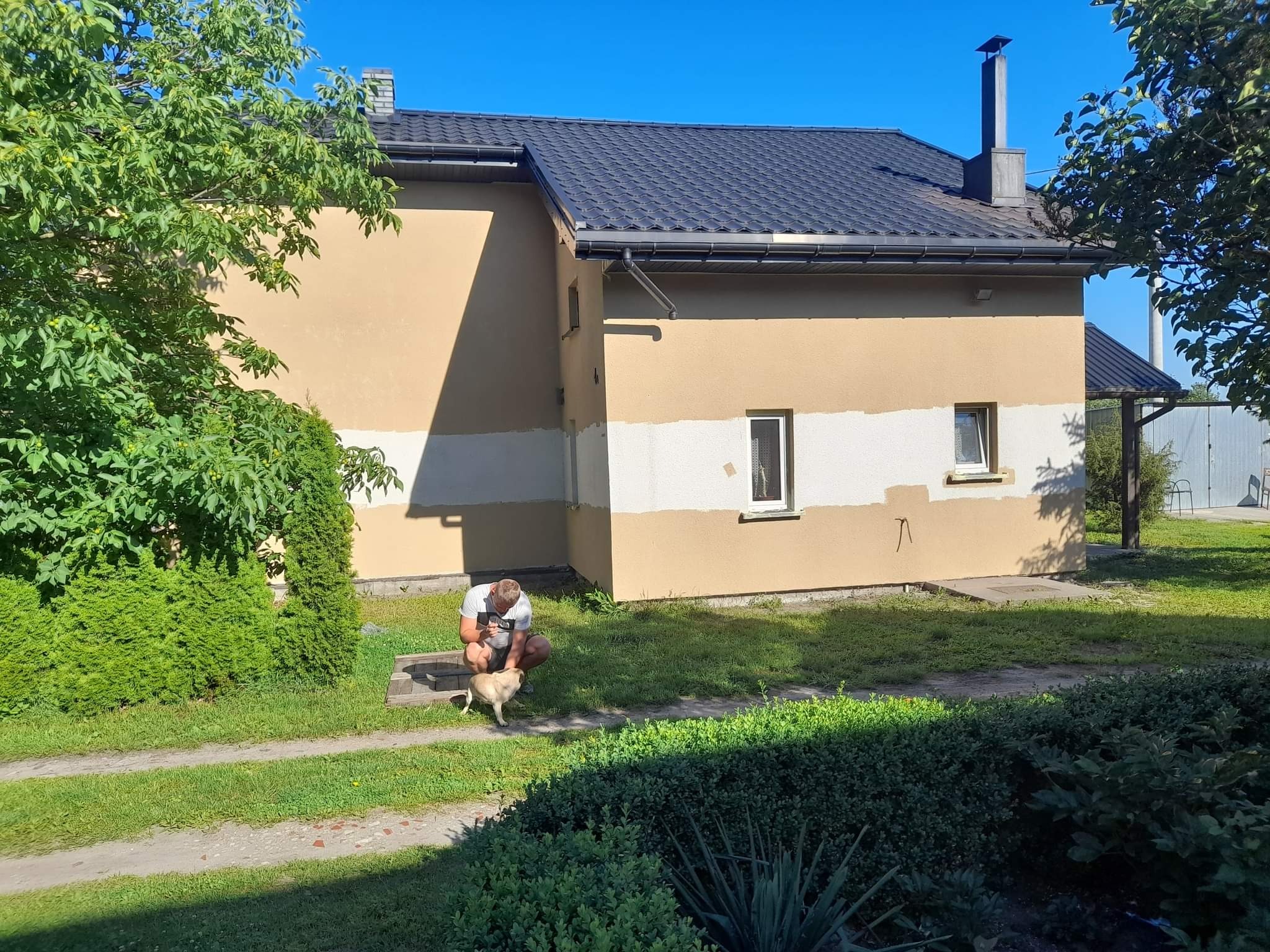 Dwukondygnacyjny dom z ciemną dachówką, otynkowany w połowie na beżowo, w połowie na biało, mężczyzna kuca obok małego psa na trawniku.