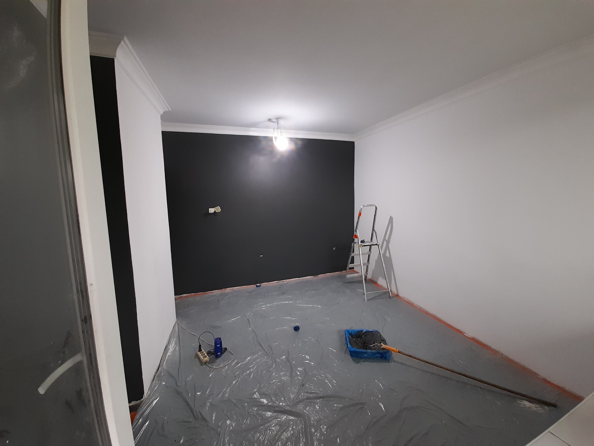 Pokój w trakcie malowania: jedna ściana pomalowana na grafitowo, pozostałe na biało, podłoga zabezpieczona folią, widoczna drabina, kuweta z farbą i przedłużka do wałka.