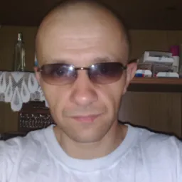 Autoportret mężczyzny w okularach przeciwsłonecznych na tle półki z różnymi przedmiotami, w tym kosmetykami i ozdobami.