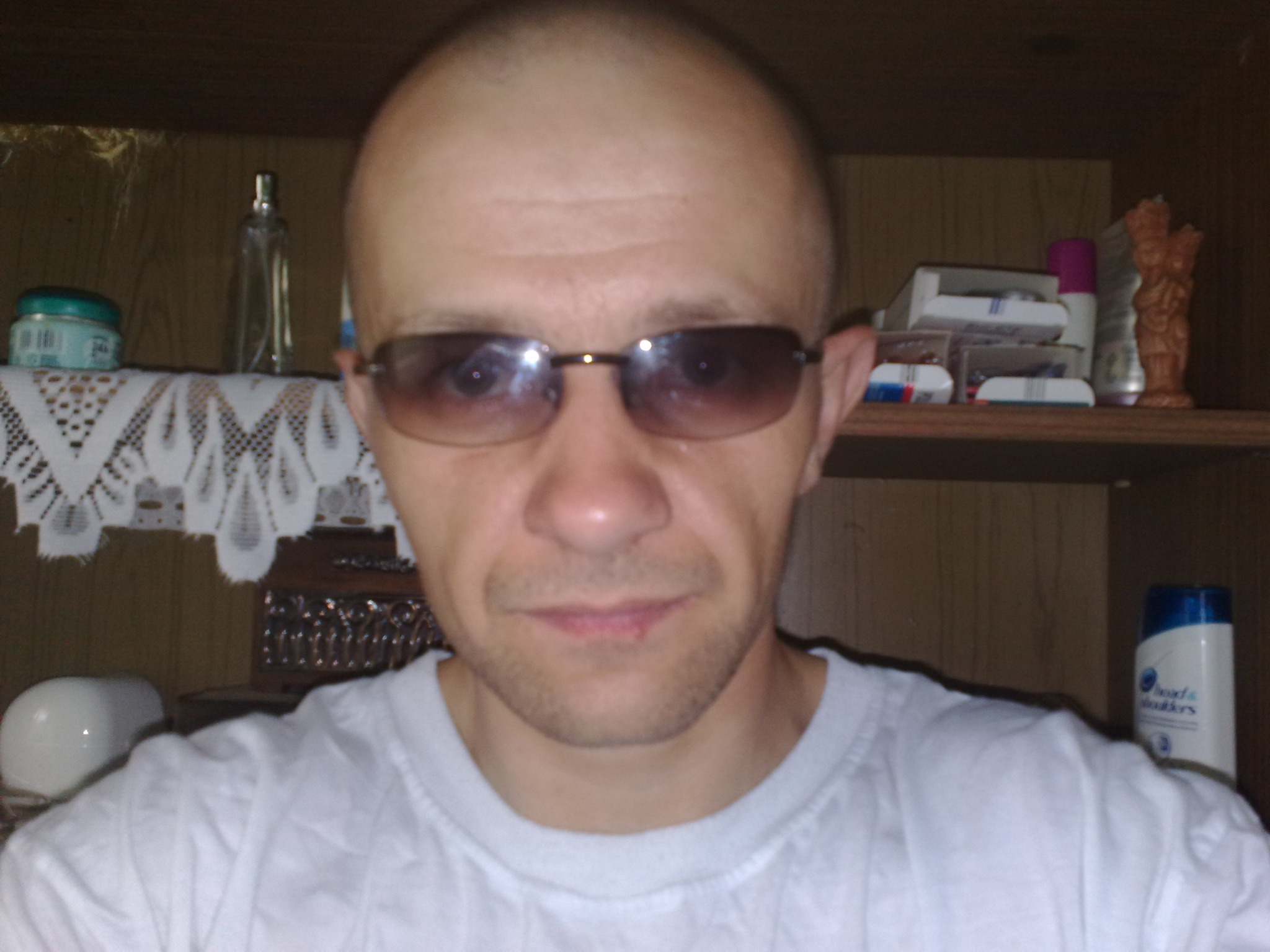 Autoportret mężczyzny w okularach przeciwsłonecznych na tle półki z różnymi przedmiotami, w tym kosmetykami i ozdobami.