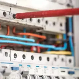 Elektryk dokręca śrubę w rozdzielnicy elektrycznej za pomocą czerwonego śrubokręta z żółtą rączką. Widoczne kable w kolorach pomarańczowym i niebieskim oraz zabezpieczenia.