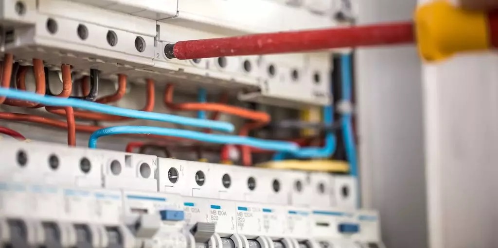 Elektryk dokręca śrubę w rozdzielnicy elektrycznej za pomocą czerwonego śrubokręta z żółtą rączką. Widoczne kable w kolorach pomarańczowym i niebieskim oraz zabezpieczenia.