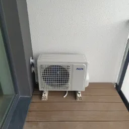 Zewnętrzna jednostka klimatyzacji marki AUX zamontowana na balkonie z drewnianą podłogą, widoczne przewody i biała ściana z fakturą.