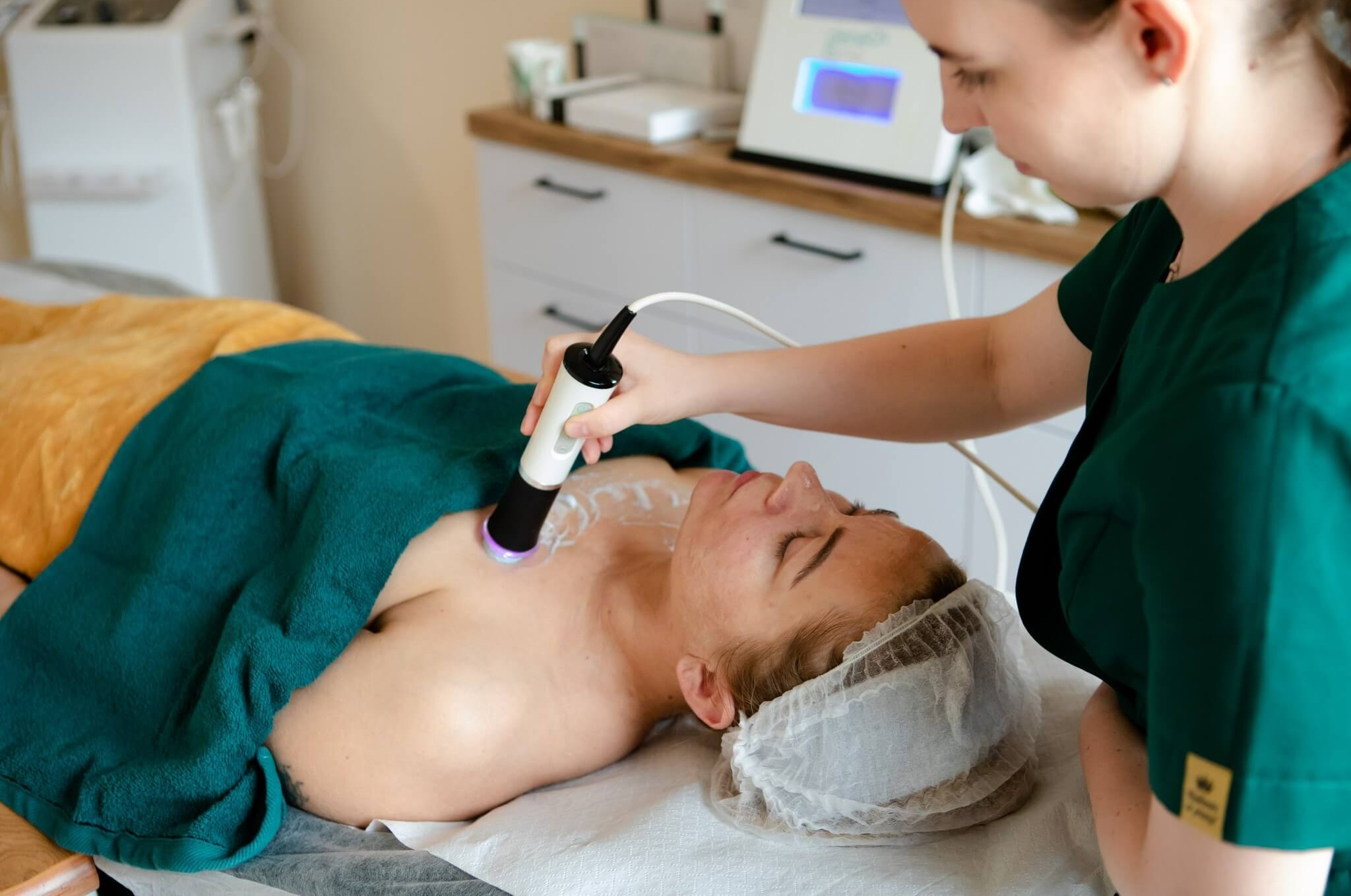 Kobieta poddawana zabiegowi na ciało z użyciem urządzenia emitującego światło, leżąca na leżance przykrytej żółtym i zielonym ręcznikiem, w tle białe meble kosmetyczne.