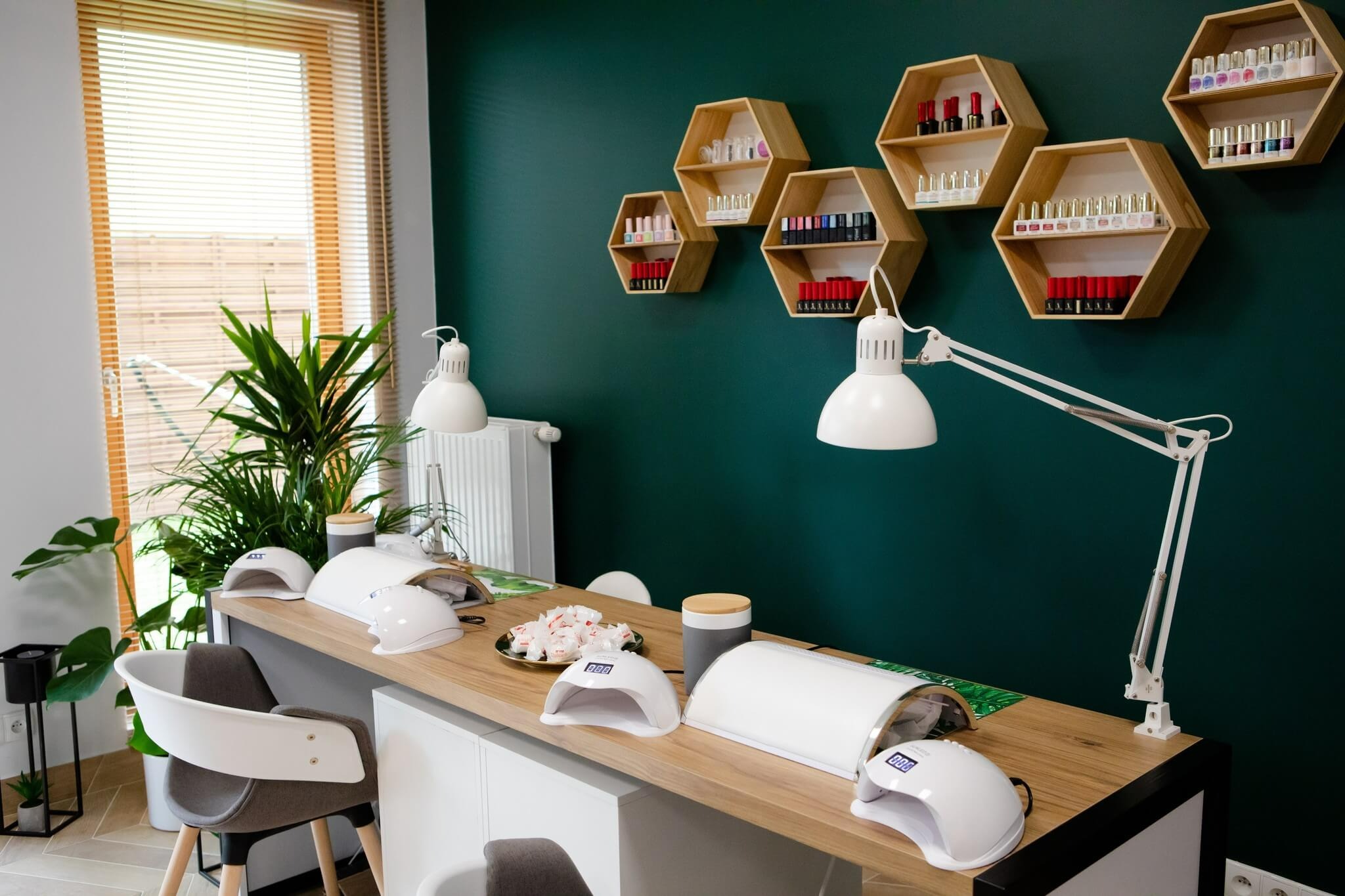 Stanowisko do stylizacji paznokci z lampami UV, pochłaniaczem pyłu, zieloną ścianą, sześciokątnymi półkami z lakierami oraz rośliną doniczkową w tle.