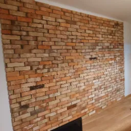 Ściana wewnętrzna wyłożona dekoracyjną cegłą w odcieniach pomarańczy i brązu, widoczna z boku, z fragmentem podłogi i mebli w tle.