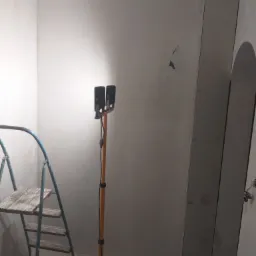 Świeżo zagruntowane ściany w trakcie remontu, oświetlone lampą na statywie, z widoczną drabiną malarską i fragmentem płyty karton-gips przy futrynie drzwiowej.