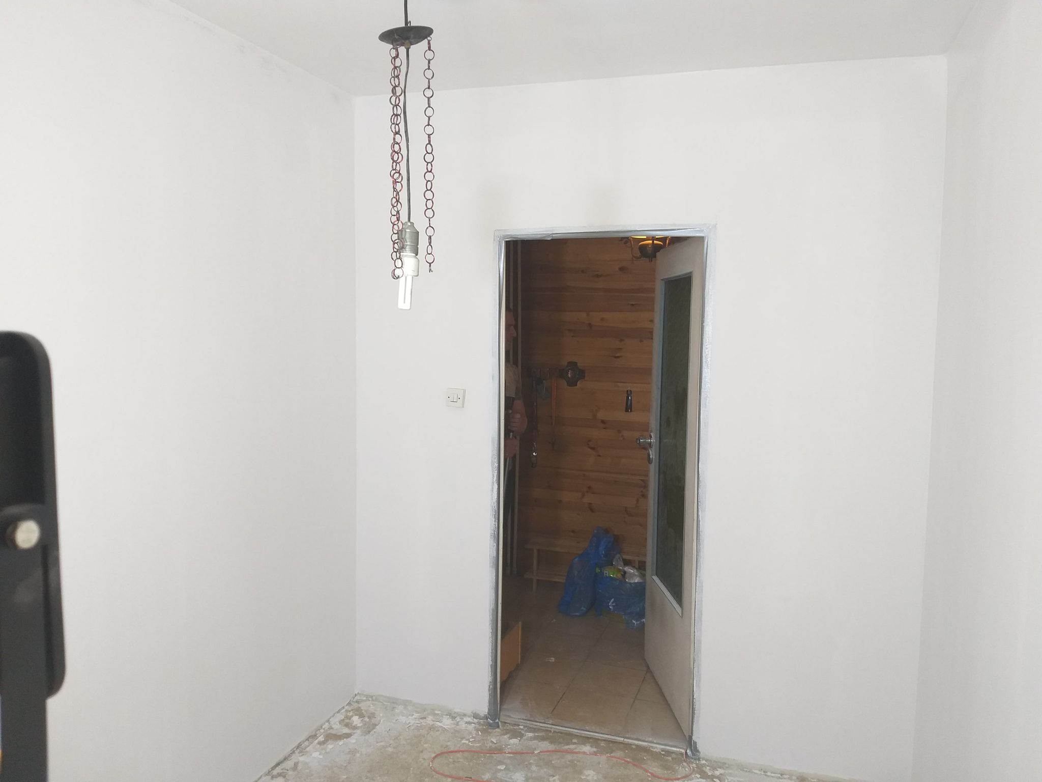 Wnętrze w trakcie remontu: świeżo pomalowane na biało ściany, surowy beton na podłodze, otwarta drewniana sauna w tle, wisząca lampa z łańcuchami.