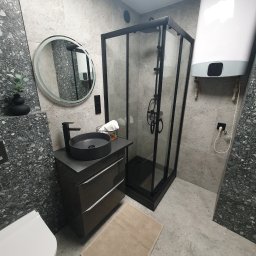 FixHome - Nowoczesna łazienka z kabiną prysznicową w czarnej ramie, okrągłym lustrem z podświetleniem LED, umywalką nablatową w kolorze grafitowym i grzejnikiem elektrycznym Ariston na ścianie wyłożonej...