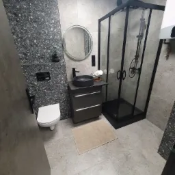 Nowoczesna łazienka z kabiną prysznicową w czarnej ramie, okrągłym lustrem nad umywalką w kolorze grafitowym, toaletą i płytkami imitującymi lastryko na ścianach.