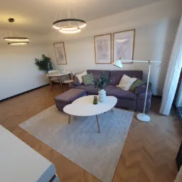 Jasne wnętrze salonu z parkietem ułożonym w jodełkę, fioletową sofą z poduszkami, białym stolikiem kawowym z wazonem, dywanem w odcieniach szarości, lampą podłogową i wiszącymi lampami w kształcie...