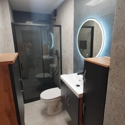 FixHome Bartosz Węglarski - Nowoczesna łazienka z szarymi płytkami, okrągłym lustrem z podświetleniem LED, czarną kabiną prysznicową i wiszącą szafką pod umywalką z drewnianym blatem.