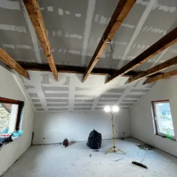 Poddasze w trakcie montażu płyt kartonowo-gipsowych, widoczne drewniane belki stropowe, tymczasowe oświetlenie halogenowe, ślady po szpachlowaniu na płytach, okna po bokach pomieszczenia.