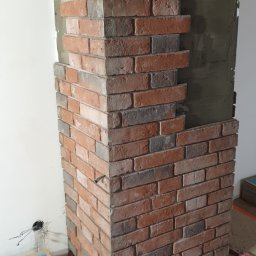 FixHome - Fragment ściany w trakcie wykańczania z cegły klinkierowej, widoczny klej na ścianie, narzędzia na podłodze, narożnik obłożony cegłą.