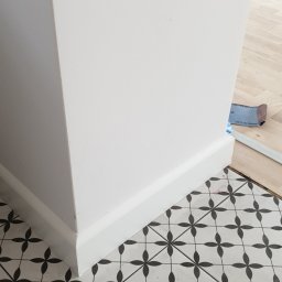 FixHome - Świeżo pomalowany narożnik ściany z białą listwą przypodłogową, stykający się z wzorzystą, czarno-białą podłogą z płytek ceramicznych; w tle widoczny fragment drewnianej podłogi zabezpieczonej...