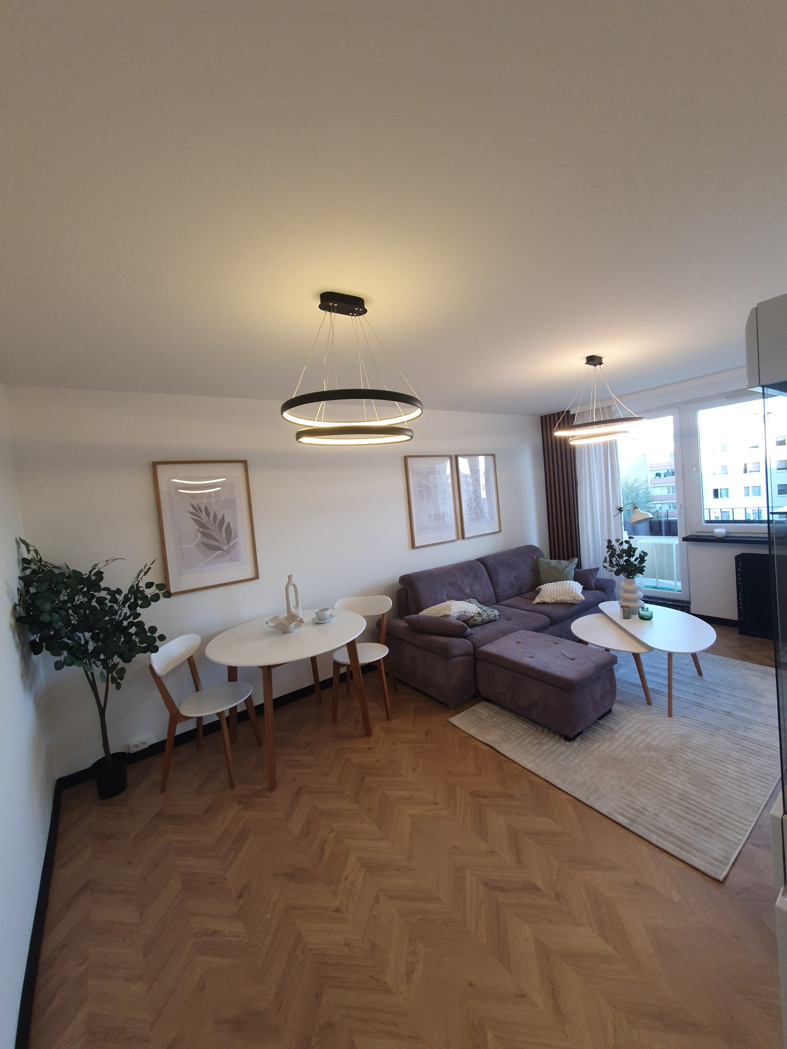 Jasne wnętrze mieszkania z parkietem w jodełkę, szarą sofą, okrągłym stołem i nowoczesnym oświetleniem, udekorowane roślinami i obrazami.