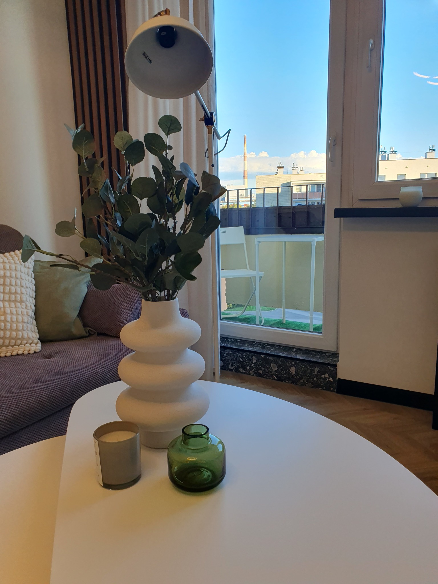 Stylowa aranżacja wnętrza z sofą, stolikiem kawowym z dekoracyjnymi wazonami i świecą, widok na balkon z roślinnością i panoramą miasta w tle.