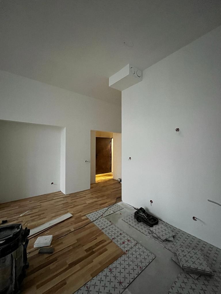 Wnętrze w trakcie remontu: częściowo ułożone panele podłogowe z drewna, fragment podłogi przygotowany pod płytki ceramiczne z motywem krzyżyków, białe ściany z widocznymi otworami po instalacjach...