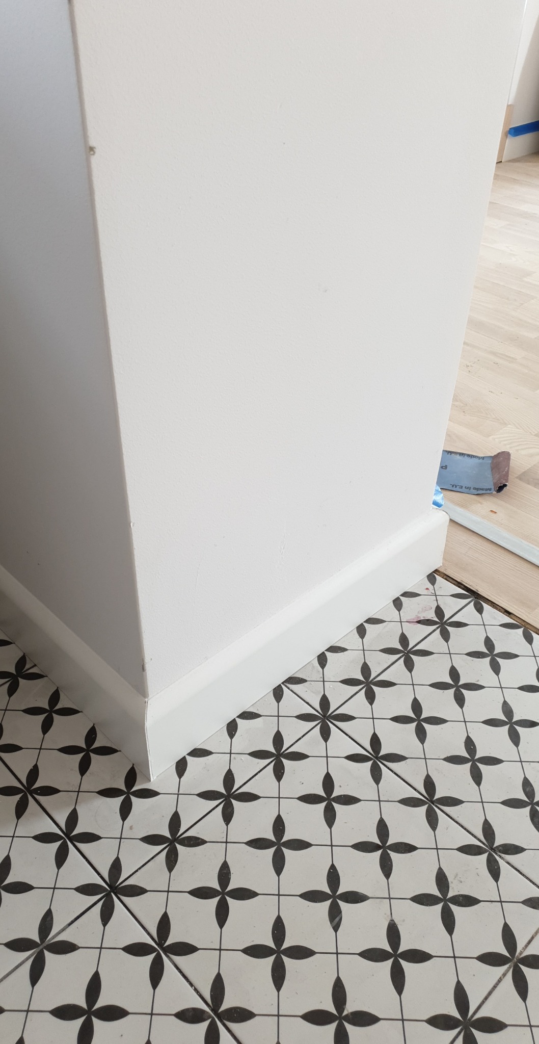 Świeżo pomalowany narożnik ściany z białą listwą przypodłogową, stykający się z wzorzystą, czarno-białą podłogą z płytek ceramicznych; w tle widoczny fragment drewnianej podłogi zabezpieczonej...