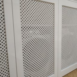 AB Meble Kamyszek Arkadiusz - Białe, lakierowane fronty meblowe z ażurową kratką, prawdopodobnie maskujące grzejnik, widoczne na tle parkietu.