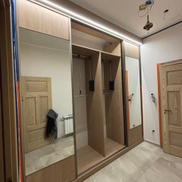 AB Meble Kamyszek Arkadiusz - Szafa wnękowa z przesuwnymi drzwiami lustrzanymi i oświetleniem LED, widoczne niedokończone prace elektryczne na suficie, odzwierciedlenie grzejnika i ubrania w lustrze.