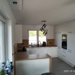 AB Meble Kamyszek Arkadiusz - Nowoczesna biała kuchnia z drewnianym blatem i wyspą, oświetlona geometryczną lampą wiszącą. Widok na okno z zielenią w tle.