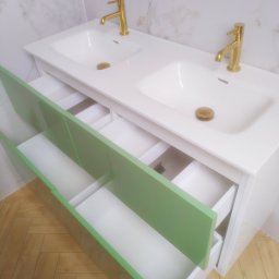 AB Meble Kamyszek Arkadiusz - Biała umywalka podwójna z otwartymi szufladami w kolorze jasnej zieleni i złotą armaturą na tle marmurowej ściany i parkietu ułożonego w jodełkę.