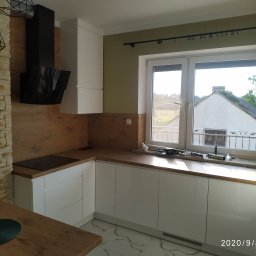 AB Meble Kamyszek Arkadiusz - Nowoczesna kuchnia z białymi szafkami i drewnianym blatem, czarny okap, widok przez okno na budynek z ogrodem, tuby z silikonem na parapecie.