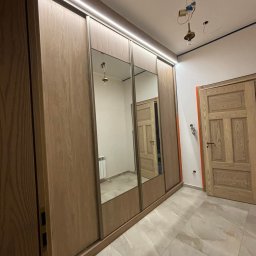 AB Meble Kamyszek Arkadiusz - Szafa wnękowa z przesuwnymi drzwiami z lustrem, oświetlenie LED, drewniane wykończenie, widoczne niedokończone prace elektryczne sufitowe i listwy przypodłogowe.