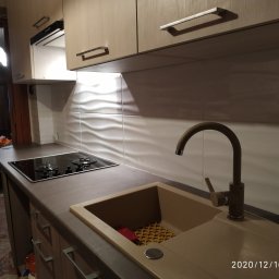 AB Meble Kamyszek Arkadiusz - Beżowa kuchnia z drewnianymi szafkami, płytą indukcyjną i zlewozmywakiem z brązową baterią, widok na salon z osobą siedzącą w fotelu.