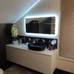 Nowoczesna łazienka z umywalką nablatową w kolorze czarnym, podświetlanym lustrem i białymi szafkami pod umywalką, uzupełniona o dekoracje i puszysty dywan.