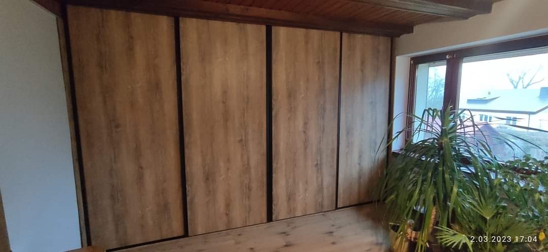 Duża szafa przesuwna z drewnopodobnymi frontami i czarnymi profilami, ustawiona pod sufitem w jasnym pokoju, z widocznym fragmentem podłogi i rośliną doniczkową przy oknie.