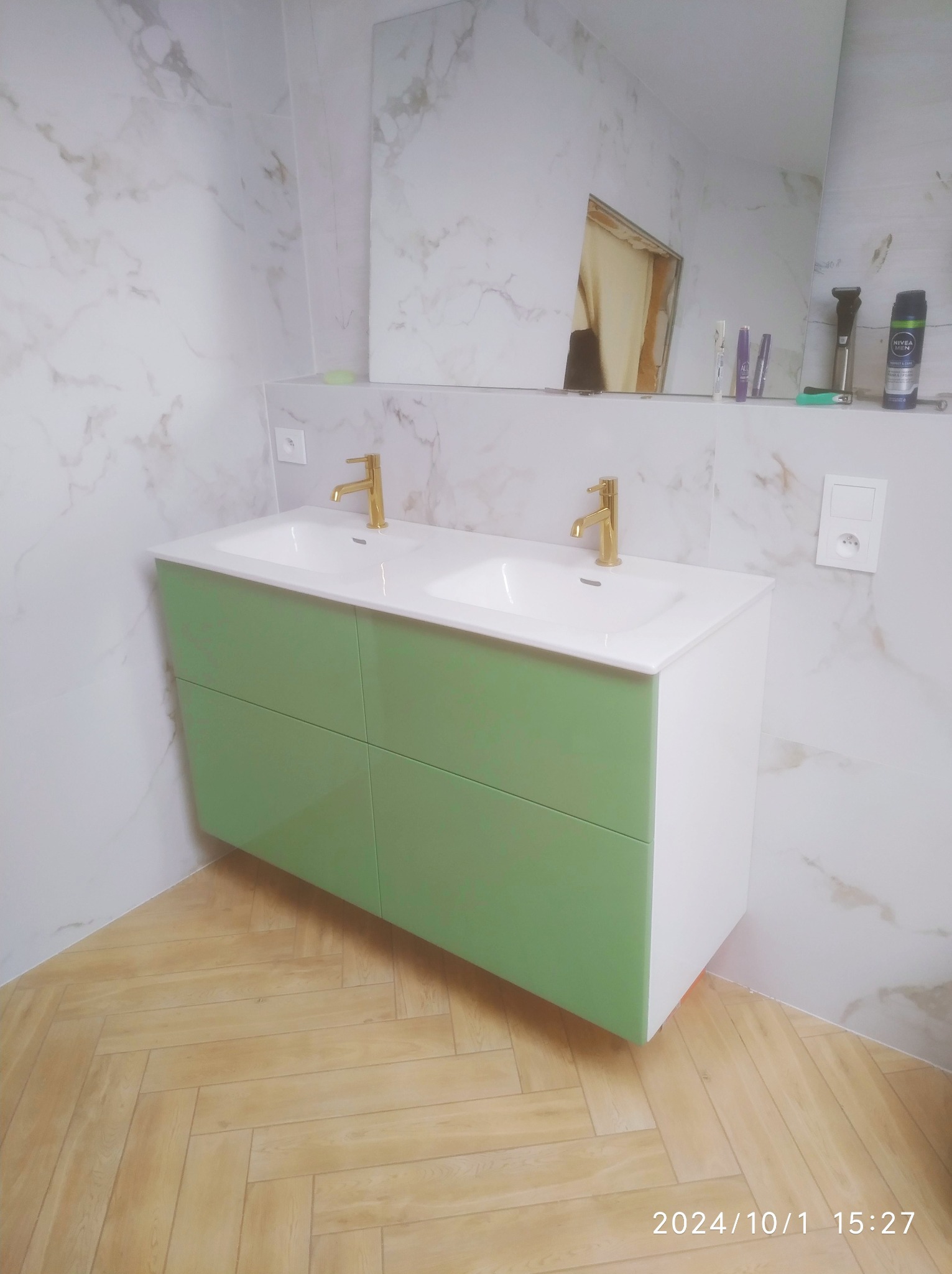 Nowoczesna łazienka z podwójną umywalką na szafce w odcieniach bieli i pastelowej zieleni, złote baterie, marmurowe płytki na ścianach i podłoga ułożona w jodełkę.