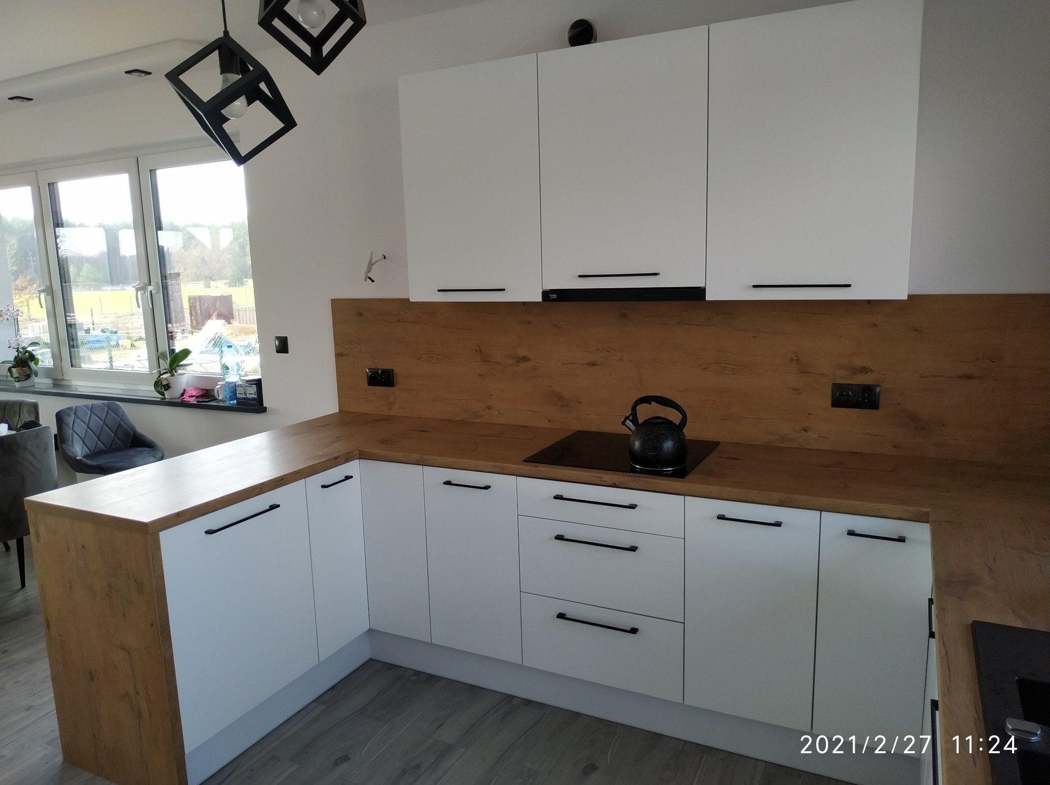 Nowoczesna kuchnia z białymi szafkami i drewnianym blatem, czajnik na płycie indukcyjnej, okno z widokiem na zewnątrz.