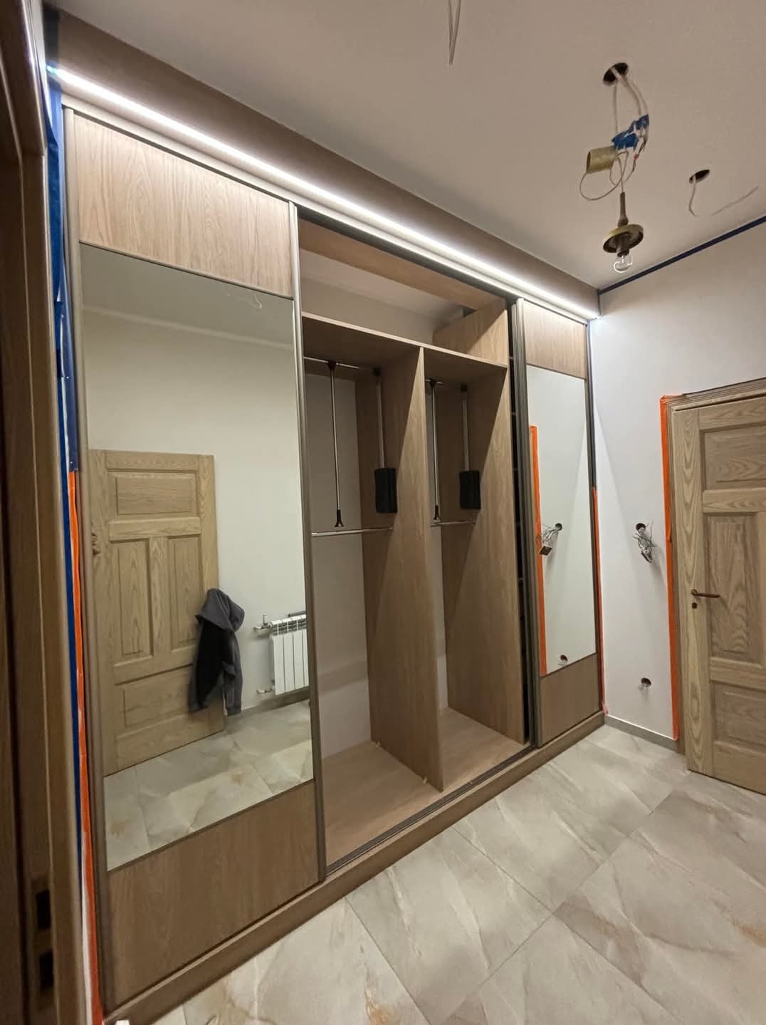 Szafa wnękowa z przesuwnymi drzwiami lustrzanymi i oświetleniem LED, widoczne niedokończone prace elektryczne na suficie, odzwierciedlenie grzejnika i ubrania w lustrze.